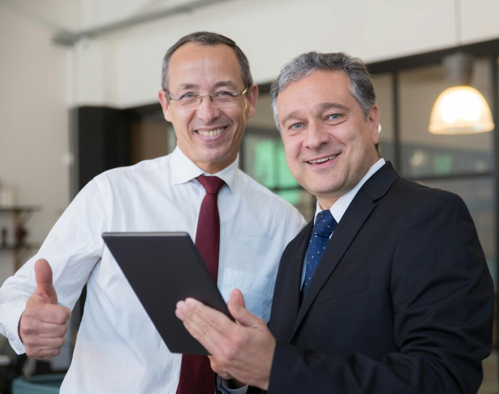 Two smiling businessmen in corporate attire discussing work on a tablet in a modern office.